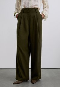 Pantalones de pierna ancha oscuros color oliva de cintura alta con pliegues frontales, combinados con una blusa crema transparente y tacones marrones de punta en un suelo gris.