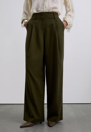 Pantalones de pierna ancha oscuros color oliva de cintura alta con pliegues frontales, combinados con una blusa crema transparente y tacones marrones de punta en un suelo gris.