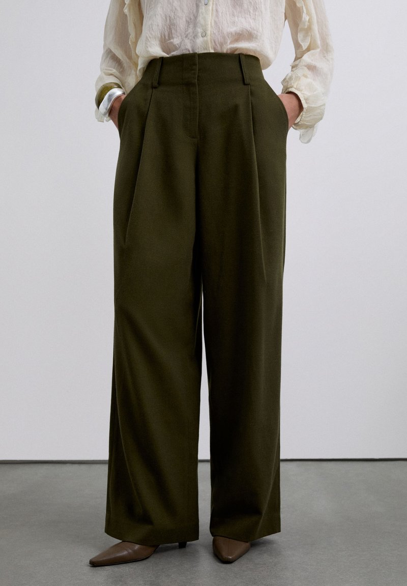 Pantalones de pierna ancha oscuros color oliva de cintura alta con pliegues frontales, combinados con una blusa crema transparente y tacones marrones de punta en un suelo gris.