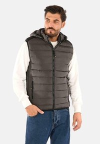 Gilet grigio imbottito con cappuccio, chiusura con zip e tasche laterali; indossato sopra una maglietta bianca a maniche lunghe. Presenta cuciture orizzontali.