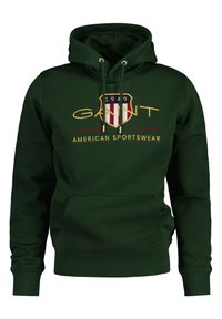 Hoodie vert foncé en tissu doux, avec une poche avant, une capuche à cordons et un emblème avec des accents rouges, blancs et dorés.