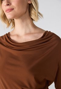 Braun drapiertes Top aus glattem Stoff mit einem Cowl-Neck-Detail, das sanfte Falten und eine lockere Passform aufweist und den Ausschnitt betont.