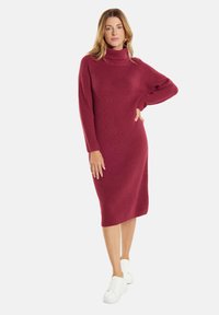 Rippstrickkleid in Bordeauxrot mit hohem Kragen und langen Ärmeln, kombiniert mit weißen Turnschuhen. Midi-Länge.