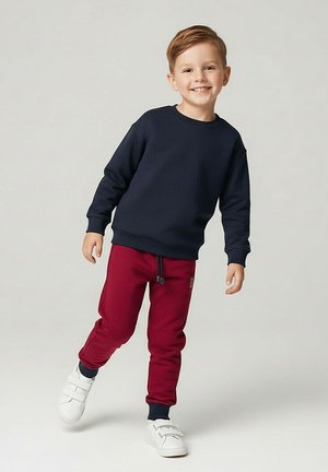 Uśmiechnięty blond chłopiec w granatowej bluzie, czerwonych spodniach dresowych i białych trampkach stoi z uniesioną jedną stopą na jasnym, jednolitym tle.