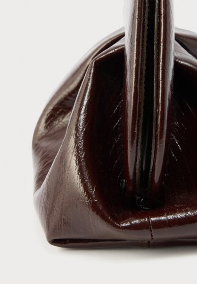 Σκούρο καφέ γυαλιστερή δερμάτινη τσάντα χειρός, εμφανιζόμενη από το πλάι, που δείχνει το δομημένο της σχήμα και την ομαλή υφή.