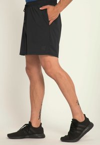 Sorte sports shorts med elastisk talje, der har sidelommer og en diskret logo på kanten. Modellen har sorte sportssko på.