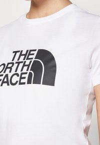 Vit t-shirt med kort ärm i bomull. Har en stor svart logotryck på framsidan med ett distinkt kurvigt designelement.