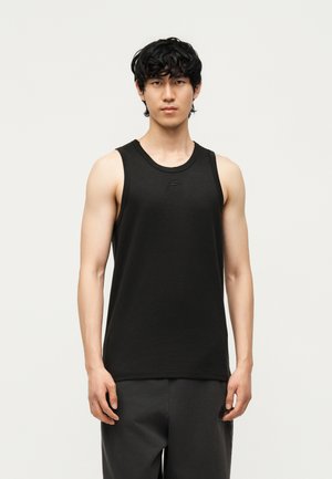 Young man with black curly hair wearing a black sleeveless tank top and dark pants, standing against a plain light background.
