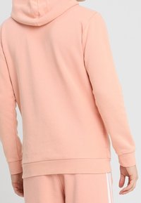 Felpa rosa con una texture morbida, dotata di tasca a marsupio, polsini e orlo a costine. Il cappuccio riposa piatto sulla schiena.