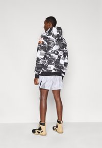 Μαύρη και λευκή φούτερ με κουκούλα και σχέδιο, με πορτοκαλί λεπτομέρειες· συνοδευόμενη από ανοιχτόγριες σορτς με γεωμετρικό σχέδιο και μαύρα αθλητικά μποτάκια.