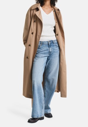 Personne portant un long trench-coat beige par-dessus un pull en maille blanc, un jean bleu clair à jambes larges et des chaussures plates noires.