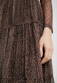 Main reposant à côté d'une robe plissée marron avec un motif de points noirs irréguliers ; manche longue transparente avec imprimé assorti visible.