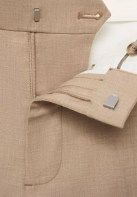 Beige dress pants close-up showing zipper, metal hook clasp, button closure, and partial view of a white shirt cuff with button.