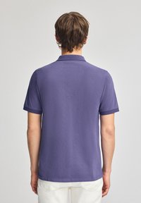 Lila pikétröja med en texturerad tygkvalitet, kort ärm och platt krage; har en rak fåll och ljusa kontrasterande detaljer.