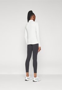 Maglietta bianca a maniche lunghe con colletto alto, abbinata a leggings grigio scuro. Il modello indossa scarpe da ginnastica grigio chiaro con accenti blu.