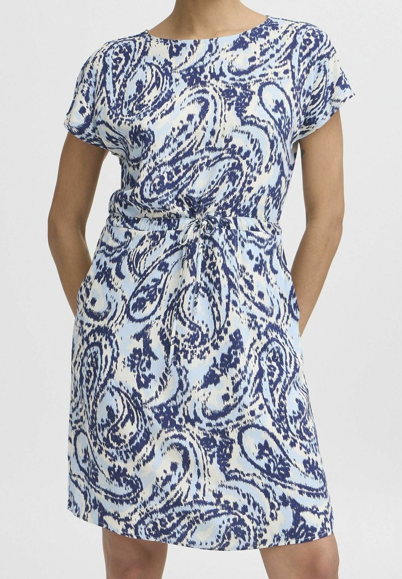 Femme portant une robe à manches courtes, longueur genou, avec un motif abstrait bleu et blanc et une ceinture nouée à la taille, mains dans les poches, sur fond blanc.