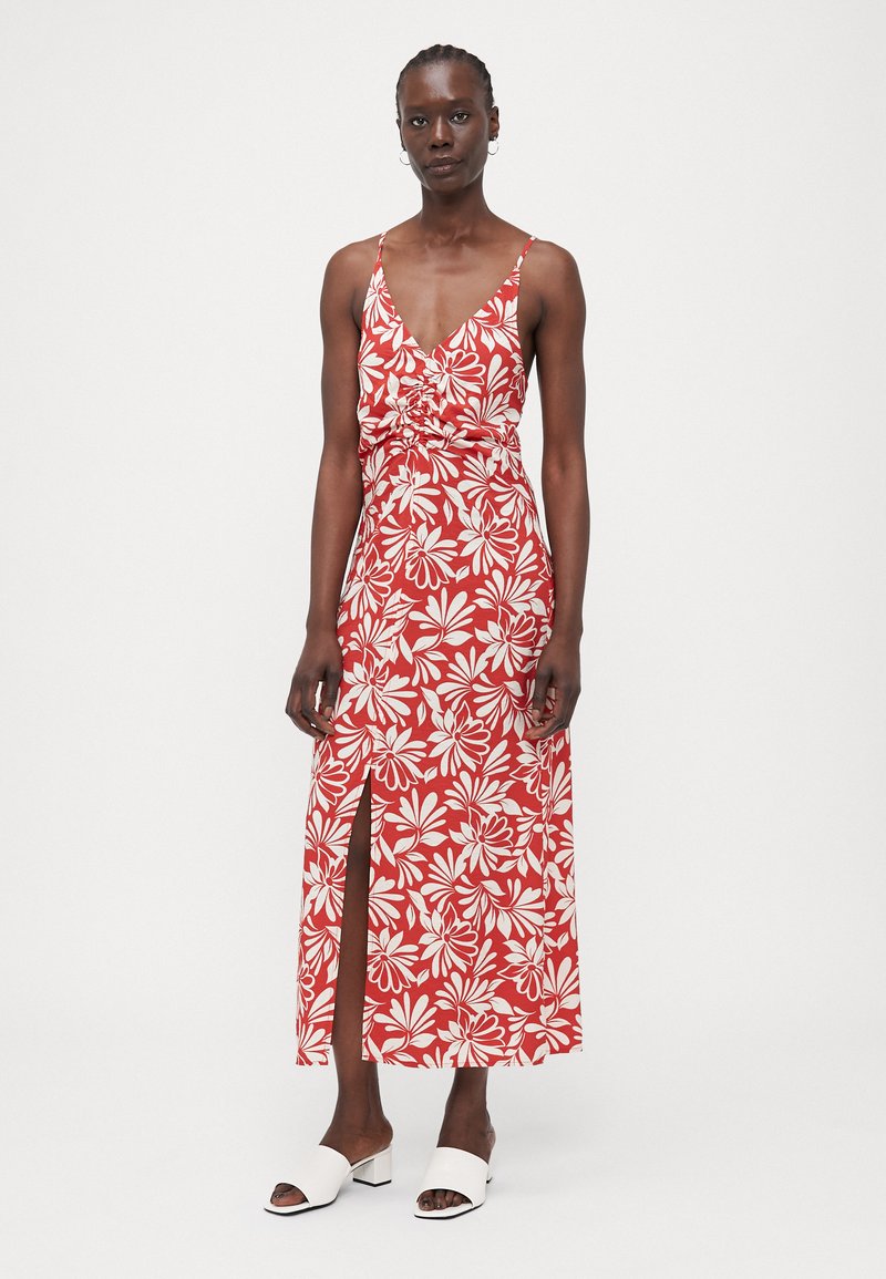 Maxikleid in Rot mit weißem Blumenmuster, ausgestattet mit einem tiefen V-Ausschnitt, verstellbaren Spaghettiträgern und einem seitlichen Schlitz. Tragen mit weißen Absatz-Mules.