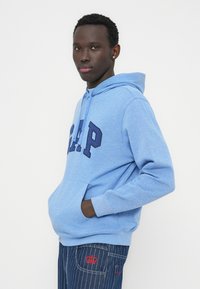 Ανοιχτό μπλε φούτερ με κουκούλα, με κεντημένα γράμματα "GAP" σε ναυτικό μπλε, ρυθμιζόμενο κορδόνι και μπροστινή τσέπη. Φορεμένο με σκουρό μπλε τζιν με ρίγες και κόκκινες λεπτομέρειες.