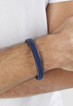 Arm mit hellbraunem Haar, der ein strukturiertes, dunkelblaues, geflochtenes Lederarmband vor weißem Ärmelfond trägt.