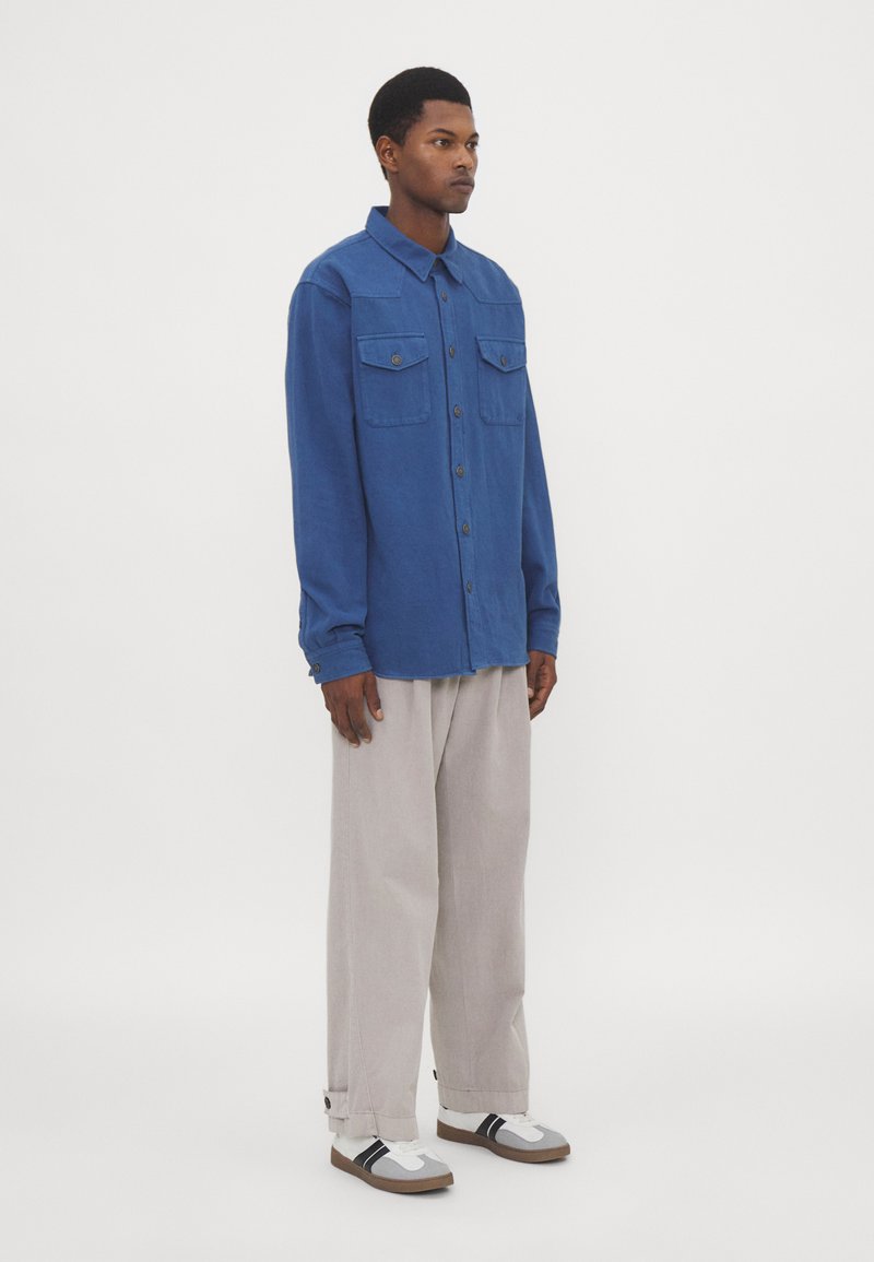 Camicia blu a maniche lunghe con bottoni e due taschini sul petto, indossata con pantaloni larghi grigio chiaro e scarpe casual. Design semplice, colore uniforme.