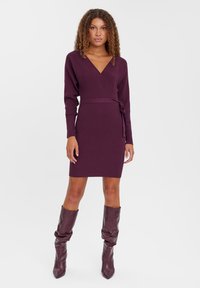 Robe portefeuille en maille de couleur violet profond avec des manches longues, décolleté en V, taille cintrée et ceinture ajustable. Portée avec des bottes marron slouchy jusqu'aux genoux.