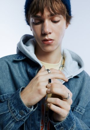 Junge Person mit einer blauen Mütze und Jeansjacke mit Kapuzenpullover, die nach unten schaut und silberne Ringe an ihren Fingern justiert.