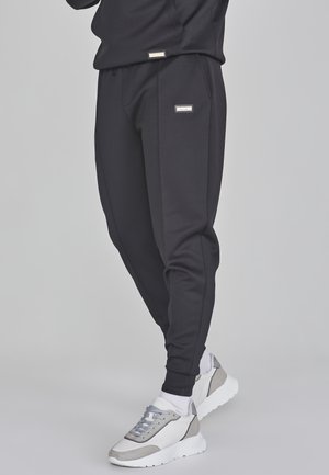 Beine in schwarzen, zulaufenden Jogginghosen mit Seitentaschen und weiß-grauen Sneakers mit weißen Socken vor einem hellgrauen Hintergrund.