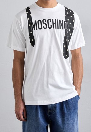 Uomo con maglietta bianca Moschino con stampa di cravatta a pois neri e jeans blu, in piedi con una mano in tasca.