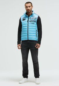 Helles blaues Pufferweste mit horizontalen Nähten, Fronttaschen und Logos. Getragen über einem schwarzen Langarmtop und kombiniert mit dunklen Jeans und weißen Sneakers.