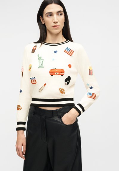 Γυναίκα που φοράει λευκό πουλόβερ με μαύρο περίγραμμα και κεντημένα εμβλήματα με θέμα τις Ηνωμένες Πολιτείες, συνδυασμένο με μαύρο δερμάτινο παντελόνι.