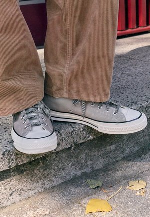 Person trägt graue Leinensneaker und braune Cordhosen und steht draußen auf einer Betontreppe mit abgefallenen Blättern in der Nähe.