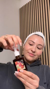 Ruskea lasipullo, jossa on valkoinen pumppukorkki ja värikäs etiketti. Sisällä oleva seerumi näyttää kirkkaalta. Käyttäjä pitää pulloa kädessä, ja kyntet ovat näkyvissä.