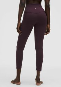 Frau, die barfuß steht und hochgeschnittene, dunkelviolette Leggings trägt, von hinten vor einem einfachen hellen Hintergrund gezeigt.