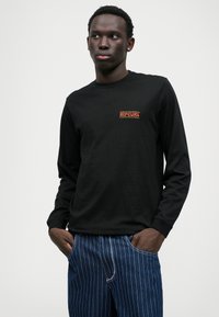 Czarna koszula z długim rękawem z małym czerwonym i żółtym logo Rip Curl na piersi, w połączeniu z niebieskimi dżinsami w paski.