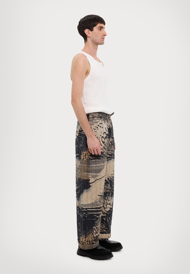 White tank top, patterned, textured, wide-legged pants in shades of beige and navy, with abstract designs; black footwear; side profile view.