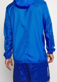 Mężczyzna w niebieskiej bluzie z kapturem i długim rękawem oraz dopasowanych niebieskich szortach z delikatnym wzorem kamuflażu, zwrócony tyłem do aparatu.