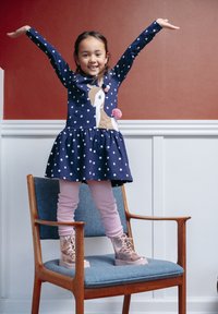 Navy Kleid mit weißen Punkten, einem Einhorn-Design und einem gestuften Rock. Helle rosa Leggings und glänzende roségoldene Stiefeletten runden das Outfit ab.
