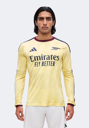 Mand iført gul langærmet Arsenal fodboldtrøje med "Emirates Fly Better" sponsorl og hvide shorts, stående mod en ensfarvet baggrund.