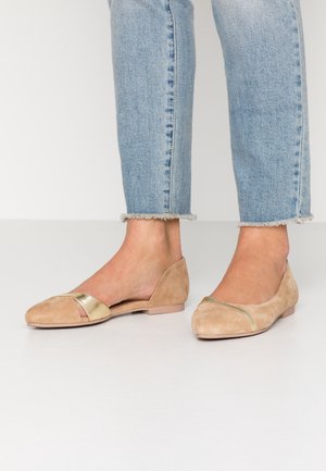 Fødder iført lyseblå jeans og beige flade sko med gyldne metalliske stropper, stående på en hvid overflade mod en ensfarvet baggrund.