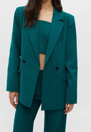 Vrouw draagt een teal dubbelgeknoopte blazer met zwarte knopen, bijpassende hooggetailleerde broek en een cropped bandeau top.