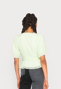 Blusa texturizada verde-claro com mangas bufantes, cintura ajustada e um cabedal fluido. Usada com calças cinza-escuro, exibindo a vista de costas.