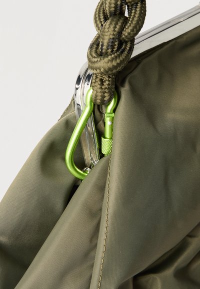 Πράσινος καρμπίνερ ασφαλισμένος σε πράσινη υφασμάτινη τσάντα με καφέ πλεγμένο σχοινί που περνά μέσα από μεταλλικό πλαίσιο.