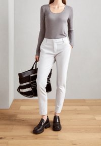 Kvinna i grå långärmad topp, vita byxor, svarta chunky loafers, håller en svart och vit randig tote-väska, står på ett trägolv.