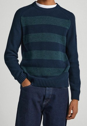 Homme portant un pull en maille rayé bleu marine et vert sur une chemise blanche avec un pantalon bleu foncé, une main dans la poche.