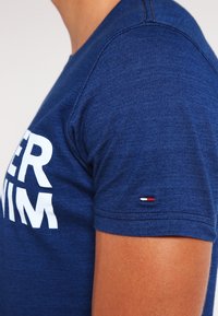 Námořnická modrá bavlněná trička s velkým bílým textem, kulatým výstřihem a krátkými rukávy. Na rukávu se nachází malá nášivka s logem v červené, bílé a modré barvě.