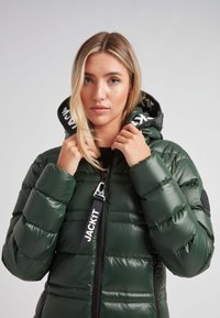 Chaqueta acolchada verde con un acabado brillante, con cierre de cremallera, capucha y acentos negros. El interior tiene un forro estampado con el nombre de la marca.