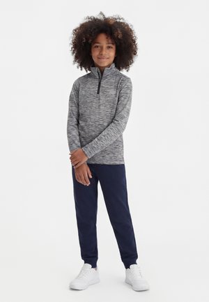 Lapsi kiharatukka seisoo harmaa vetoketjupoolover yllään, päällä tummansiniset verkkarit ja valkoiset lenkkarit valkoista taustaa vasten.