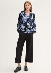 Blusa svasata nera con motivi astratti viola e blu, abbinata a pantaloni neri a gamba larga e mocassini neri.