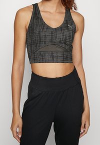 Szara sportowa biustonosz z efektem tie-dye, z głębokim dekoltem w kształcie litery V i wycięciem w talii. Wykonana z elastycznego materiału. Połączona z czarnymi spodniami z wysokim stanem.