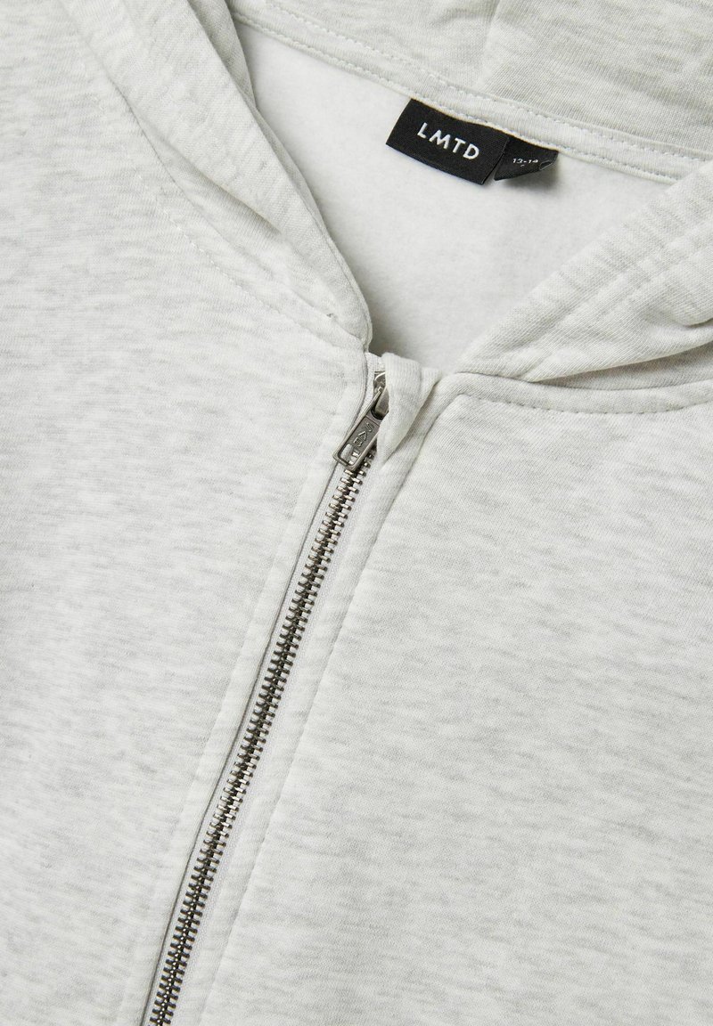 Lichtgrijze rits-hybride hoodie van zachte stof, met een capuchon, metalen rits en een label met de tekst "LMTD" binnen de halslijn.